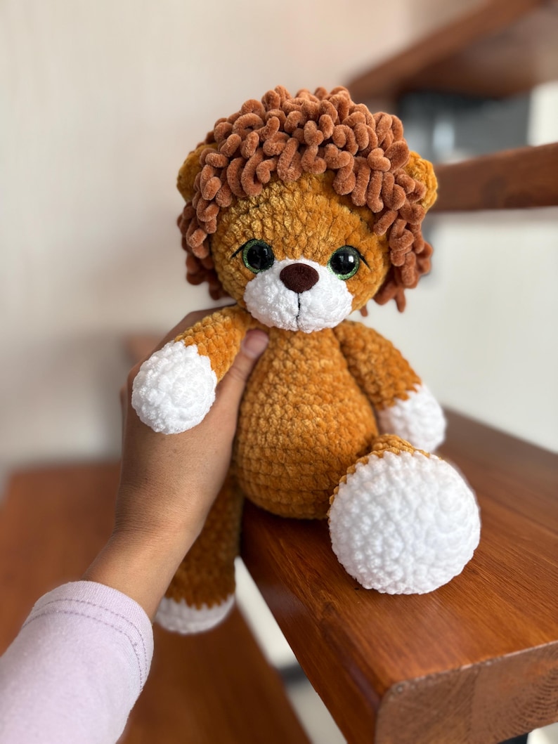 May include: A handmade crochet lion plush toy. The lion has a brown mane, a tan body, and white accents on its paws and muzzle. It has large, expressive eyes and a cute, friendly expression. The plush is soft and cuddly.