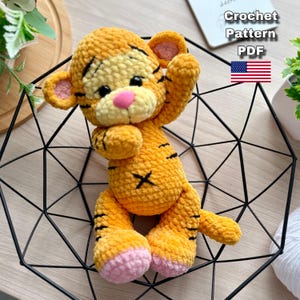 May include: A handmade orange and yellow crochet tiger toy with black stripes and a pink nose and feet. The toy is in a black geometric metal basket. Text on the image reads "Crochet Pattern PDF" with a US flag.