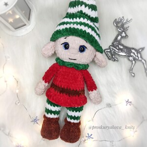 May include: A handmade crochet elf doll with a green and white striped hat, red tunic, and green and white striped pants. The doll has brown boots and a green collar. The doll's face has blue eyes and a neutral expression.