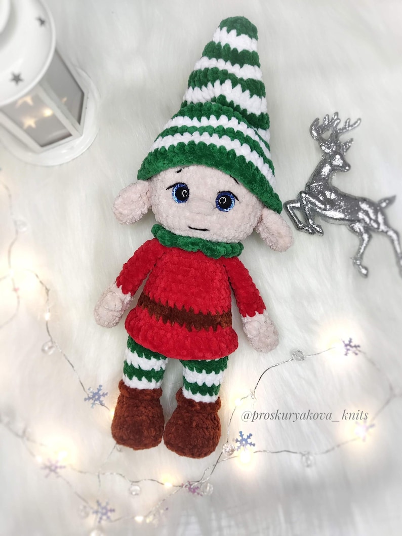 May include: A handmade crochet elf doll with a green and white striped hat, red tunic, and green and white striped pants. The doll has brown boots and a green collar. The doll's face has blue eyes and a neutral expression.