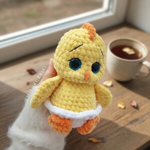 Pode incluir: Um pintinho de croch&ecirc; amarelo feito &agrave; m&atilde;o com olhos azuis, p&eacute;s laranja e uma fralda branca. O pintinho tem uma pequena crista laranja e est&aacute; sobre uma superf&iacute;cie de madeira. Uma x&iacute;cara de ch&aacute; est&aacute; ao fundo.