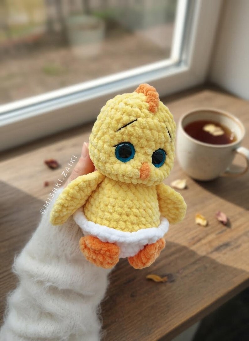 Pode incluir: Um pintinho de croch&ecirc; amarelo feito &agrave; m&atilde;o com olhos azuis, p&eacute;s laranja e uma fralda branca. O pintinho tem uma pequena crista laranja e est&aacute; sobre uma superf&iacute;cie de madeira. Uma x&iacute;cara de ch&aacute; est&aacute; ao fundo.
