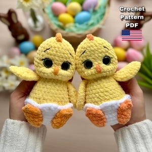 Peut inclure: Deux poussins crochetés à la main, jaunes, avec des pieds et des becs oranges, et des couches blanches. Les poussins ont de grands yeux noirs et sont tenus devant un arrière-plan flou avec des œufs de Pâques et un panier. Le texte "Crochet Pattern PDF" est inclus.