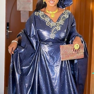 May include: A woman wearing a navy blue, flowing dress with gold embroidery around the neckline. She is also wearing a matching head wrap, gold jewelry, and carrying a red and gold clutch. The dress has a subtle dotted pattern.