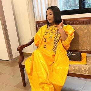 May include: A woman wearing a bright yellow, flowing dress with intricate geometric patterns on the bodice. The dress has long sleeves and a matching head covering. She is seated on a wooden bench with a patterned cushion, with a small brown purse.