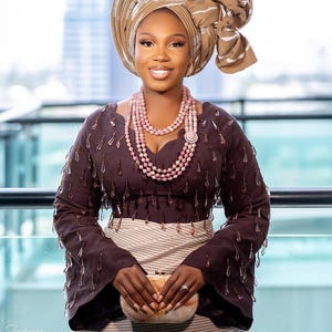 May include: A woman wearing a brown traditional outfit with a matching head wrap and a pink beaded necklace. The long-sleeved top has decorative embellishments. She is holding a small, light-colored clutch.