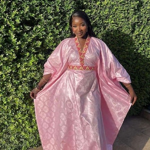 May include: A light pink, flowing dress with a floral pattern. The dress features a V-neckline and a decorative band around the waist and neckline with red and gold accents. The sleeves are wide and the dress is floor-length.