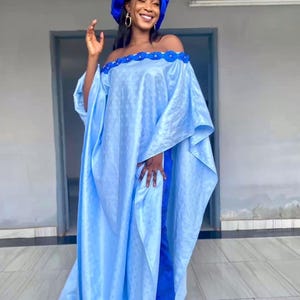 May include: A light blue off-the-shoulder dress with a matching head wrap. The dress features a floral trim along the neckline and a flowing, loose fit. The outfit is completed with blue sandals.