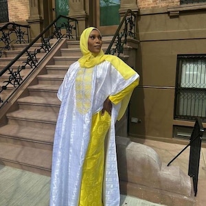 May include: A woman wearing a yellow hijab and a white and yellow outfit. The white outer garment has intricate embroidery on the front. The outfit is worn outdoors, with a brick building and stairs in the background.