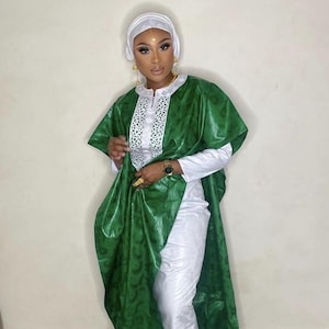 May include: A green and white ensemble featuring a long, flowing green overcoat with a subtle pattern. The outfit includes a white long-sleeved top and matching pants. The ensemble is complemented by a white head covering and gold jewelry.