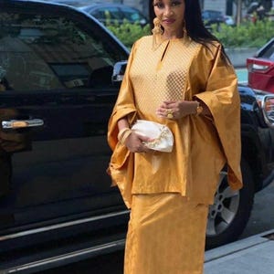 May include: A woman wearing a mustard yellow ensemble, including a long-sleeved top and a matching skirt. The outfit features a textured pattern on the bodice and skirt. She is holding a white clutch with a gold chain and wearing gold jewelry.