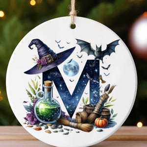 May include: A round white ceramic ornament featuring a large letter M with a dark blue starry night sky pattern. The design includes a purple witch hat, a green potion bottle, a broom, a small orange pumpkin, and a flying bat illustration.