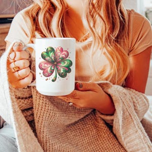 May include: White ceramic mug featuring a colorful four-leaf clover design in shades of pink, green, and gold. The clover is the focal point, surrounded by small star accents. The mug has a large handle.