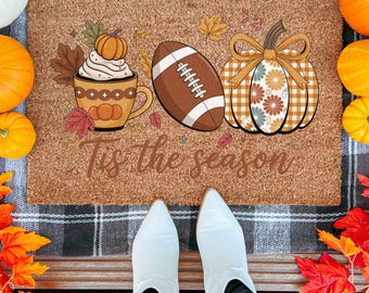 May include: A rectangular doormat with a fall theme. It features a coffee cup with whipped cream, a football, and a pumpkin with a bow. The text "Tis the season" is printed on the mat. Pumpkins and fall leaves surround the mat.