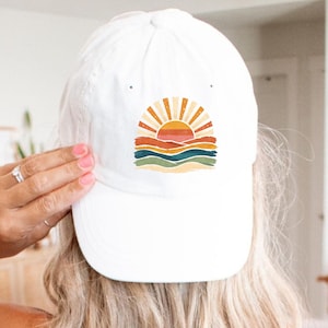 May include: A white baseball cap featuring a graphic print of a stylized sun rising over layered hills. The design includes warm tones of orange, yellow, and red, contrasted with wavy bands of teal and sage green, all centered on the front panel.