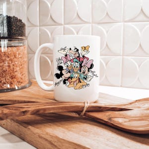 Puede incluir: Taza de cer&aacute;mica blanca con un gr&aacute;fico colorido de personajes de Disney, incluyendo Mickey Mouse, Minnie Mouse, Donald Duck, Daisy Duck, Goofy y Pluto. La taza tiene un asa grande y est&aacute; sobre una superficie de madera.