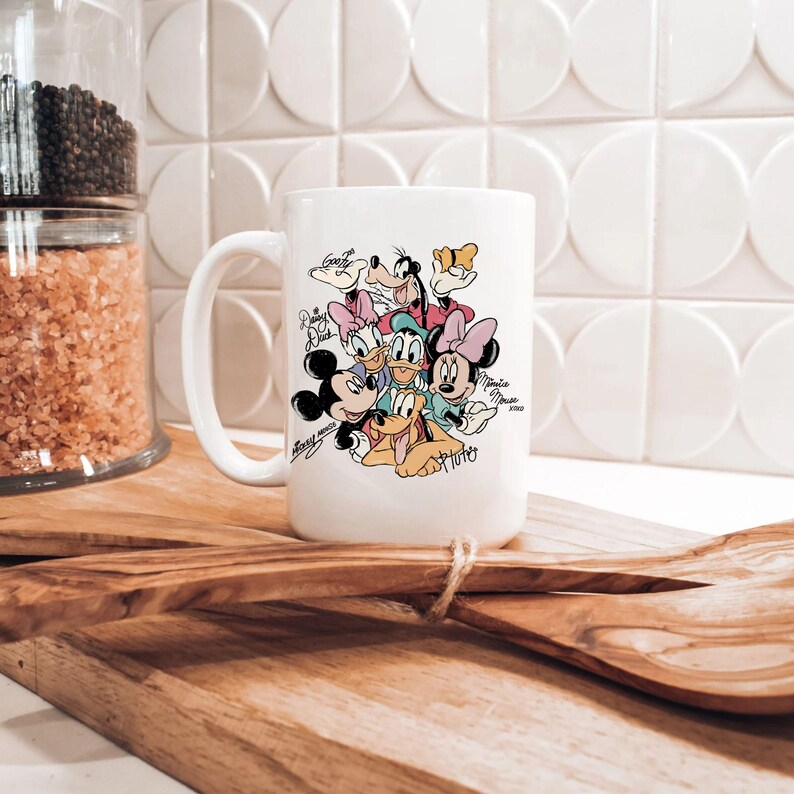 Puede incluir: Taza de cer&aacute;mica blanca con un gr&aacute;fico colorido de personajes de Disney, incluyendo Mickey Mouse, Minnie Mouse, Donald Duck, Daisy Duck, Goofy y Pluto. La taza tiene un asa grande y est&aacute; sobre una superficie de madera.