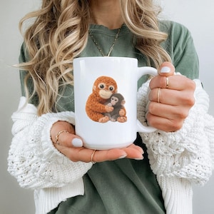 May include: White ceramic mug featuring a detailed illustration of two monkeys, one embracing the other. The larger monkey is orange, and the smaller one is dark brown. The mug has a large handle and a smooth, glossy finish.