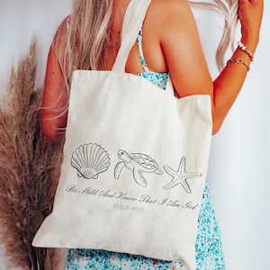 May include: Cream-colored tote bag featuring line drawings of a seashell, turtle, and starfish. The text "Be Still And Know That I Am God" and "Psalm 46:10" are printed below the illustrations. The bag has two handles.