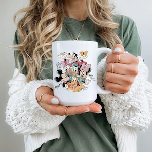 Puede incluir: Taza de cer&aacute;mica blanca con una colorida ilustraci&oacute;n de personajes de Disney, incluyendo Mickey Mouse, Minnie Mouse, Donald Duck y Pluto. La taza tiene un asa grande y firmas de personajes.