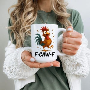 May include: White ceramic mug featuring a cartoon rooster with a red comb, brown body, and green tail. The mug has the text "F.CAW-F" below the rooster. The mug is held by a person.