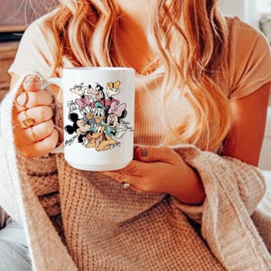 Puede incluir: Taza de cer&aacute;mica blanca con un gr&aacute;fico colorido de personajes de Disney, incluyendo Mickey Mouse, Minnie Mouse, Donald Duck y Goofy. La taza tiene un asa grande y es sostenida por una persona.