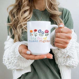 May include: White ceramic mug with the text "big fan of human rights" in colorful letters. The mug features a floral design with orange, red, blue, and pink flowers. The mug is held by a person wearing a green shirt and a white sweater.