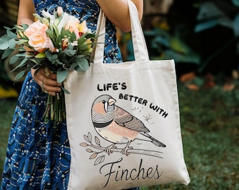 May include: A cream-colored tote bag with the text "Life's Better With Finches" and a graphic of a finch perched on a branch. The finch has a pink breast and gray head. The bag has two handles.