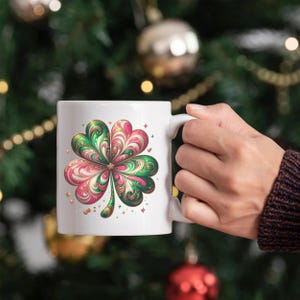 May include: White ceramic mug featuring a colorful four-leaf clover design. The clover has green, pink, and gold accents, with small star details. The mug is held by a person, with a blurred background of Christmas decorations.