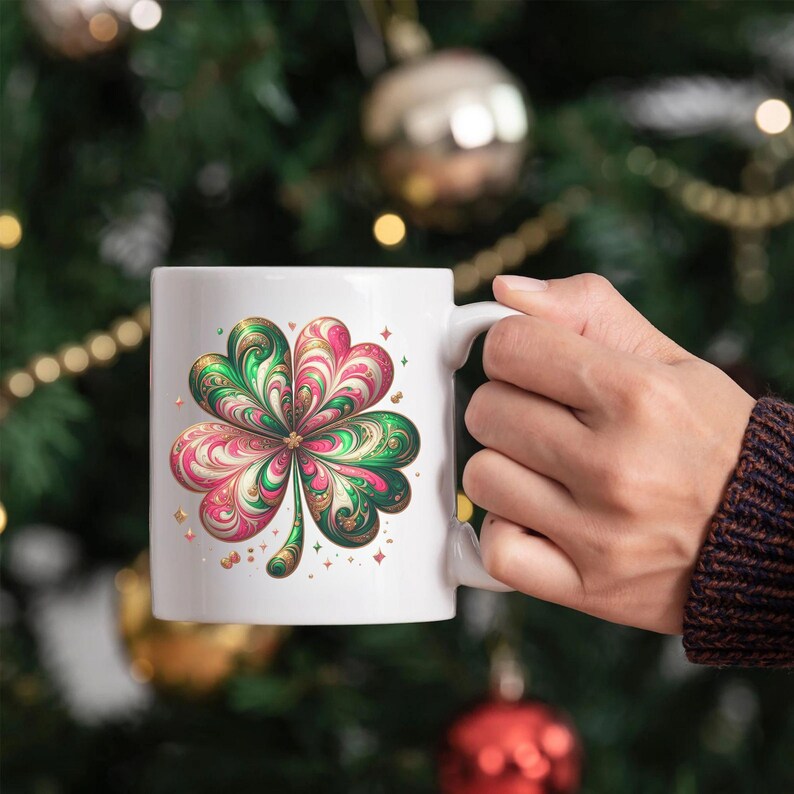 May include: White ceramic mug featuring a colorful four-leaf clover design. The clover has green, pink, and gold accents, with small star details. The mug is held by a person, with a blurred background of Christmas decorations.