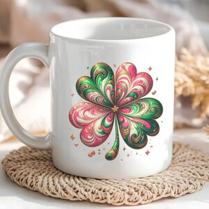 May include: White ceramic mug featuring a colorful four-leaf clover design. The clover has green, pink, and gold accents with a swirling pattern. Small gold stars and shapes surround the clover. The mug sits on a woven coaster.