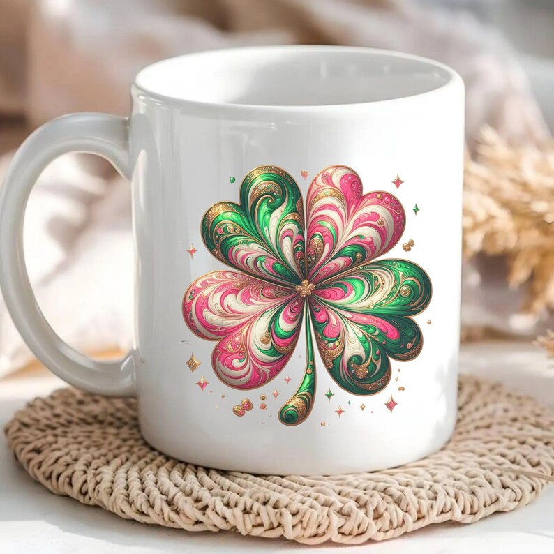 May include: White ceramic mug featuring a colorful four-leaf clover design. The clover has green, pink, and gold accents with a swirling pattern. Small gold stars and shapes surround the clover. The mug sits on a woven coaster.
