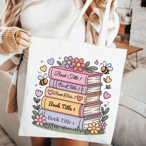 May include: A white canvas tote bag featuring a colorful graphic of a stack of five books. Each book spine is labeled with the text Book Title 1. The illustration includes pastel-colored flowers, hearts, and two small bees surrounding the books.