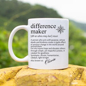May include: A white ceramic mug featuring black text that defines the term difference maker, including a phonetic pronunciation, a descriptive paragraph, synonyms, and the name Olivia, accompanied by a small black compass rose graphic.