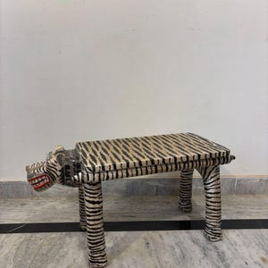May include: A decorative bench with a tiger-like design. The bench features a rectangular seat and four legs, all adorned with black and white stripes. The front of the bench is sculpted to resemble a tiger's head, complete with a red mouth.