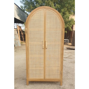 May include: A light brown wooden cabinet with a woven rattan door design. The cabinet has an arched top and two doors with vertical handles. The cabinet is standing on a dirt surface.