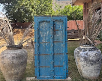 Pode incluir: Uma porta de madeira azul desgastada com ferragens metálicas está em pé sobre uma área gramada. Dois vasos cinzentos grandes e envelhecidos, cada um cheio de gramíneas ornamentais secas, ladeiam a porta. A cena é ao ar livre, com um edifício e árvores ao fundo.