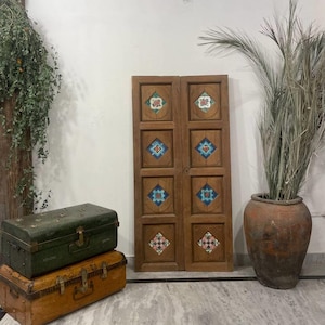 May include: Two wooden doors with inlaid, colorful square tile designs. The doors are flanked by a large terracotta pot with dried plants and two antique trunks, one green and one tan. The doors are a focal point.