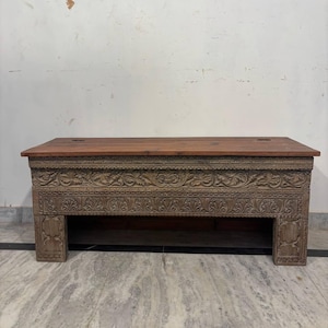 May include: A rectangular wooden coffee table with a reddish-brown top and intricately carved floral patterns on the front and sides. The table has a lower shelf and is supported by four sturdy legs. The dimensions are approximately 48 inches long, 20 inches wide, and 18 inches high.