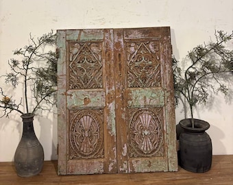 May include: A pair of weathered, intricately carved wooden doors with a distressed finish. The doors feature detailed floral and geometric patterns, with remnants of faded green and pink paint. The doors are propped up on a wooden surface, with decorative vases and branches on either side.