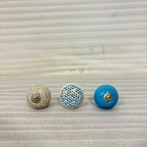 May include: Three decorative cabinet knobs. One is beige with a crackle finish, the second has a geometric pattern of black, blue, and white triangles, and the third is a solid turquoise blue. Each knob has a metal base.