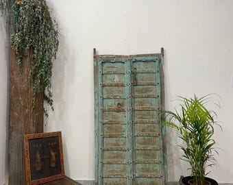 May include: An antique, weathered teal door with metal accents stands against a white wall. A small wooden stool with a framed artwork sits to the left. A tall wooden post with greenery and a potted plant complete the scene.