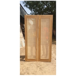 May include: Two wooden doors with woven cane inserts. The doors are a light brown color and appear to be new. The doors are standing upright on a sandy surface. The doors are framed with a light brown wood.