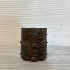 Op de afbeelding: Een cilindrische, antieke houten container met ingewikkelde gravures en metalen accenten. De donkerbruine container heeft decoratieve banden, een centrale handgreep en sierlijke bloempatronen, wat wijst op een vintage of handgemaakte oorsprong.