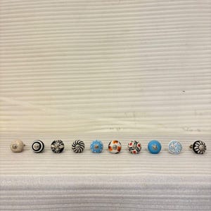 Könnte beinhalten: Eine Sammlung dekorativer Schrankknöpfe. Die Knöpfe weisen verschiedene Designs auf, darunter Blumenmuster, geometrische Formen und unifarbene Farben. Die Knöpfe sind in einer Reihe angeordnet und zeigen ihre vielfältigen Stile und Farben.