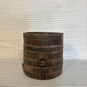 Peut inclure: Un récipient cylindrique en bois avec des bandes métalliques et une poignée en anneau métallique. Le récipient a un aspect rustique et vieilli, avec un grain de bois visible et des tons bruns foncés. Les bandes sont rivetées au bois.