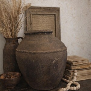 Könnte beinhalten: Ein rustikales Stillleben mit einem großen, strukturierten braunen Steinguttopf. Eine kleinere Vase mit getrocknetem Weizen, eine Holzschale, ein Stapel alter Bücher und eine Perlenkette mit Quaste vervollständigen die Anordnung. Die Gegenstände stehen auf einer dunklen Holzoberfläche.