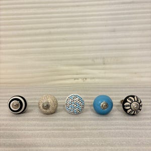 以下が含まれることがあります： 5つの装飾的なキャビネットノブ。ノブは丸く、白と黒のストライプ、ひび割れ仕上げ、青とグレーの幾何学模様、無地のライトブルー、白と黒の花柄など、さまざまなデザインが施されています。各ノブには金属製のフィッティングが付いています。
