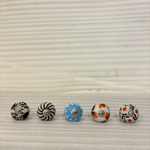 May include: Five decorative ceramic cabinet knobs. The knobs feature various designs, including black and white floral patterns, a blue knob with white dots, and knobs with orange flowers and red berries. Each knob has a silver metal base.