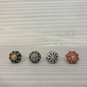 Könnte beinhalten: Vier dekorative Keramik-Schrankknöpfe mit floralen Designs. Die Knöpfe weisen eine Vielzahl von Farben auf, darunter Blau, Orange, Rot und Gold. Jeder Knopf hat eine Metallbasis zur Befestigung.